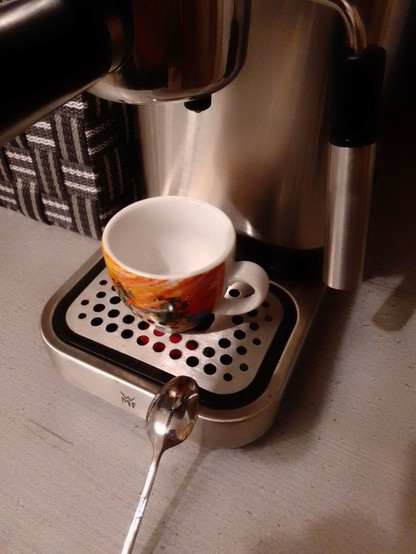 Espresso-Tasse unter dem Sieb-träger einer Espressomaschine. Das Sieb ist mit der Maximalmenge an Kaffeemehl für zwei Tassen beladen. In der Tasse befindet sich bereits der Zucker für einen sehr starken doppelten Espresso. Ein Löffel zum Umrühren liegt bereit.